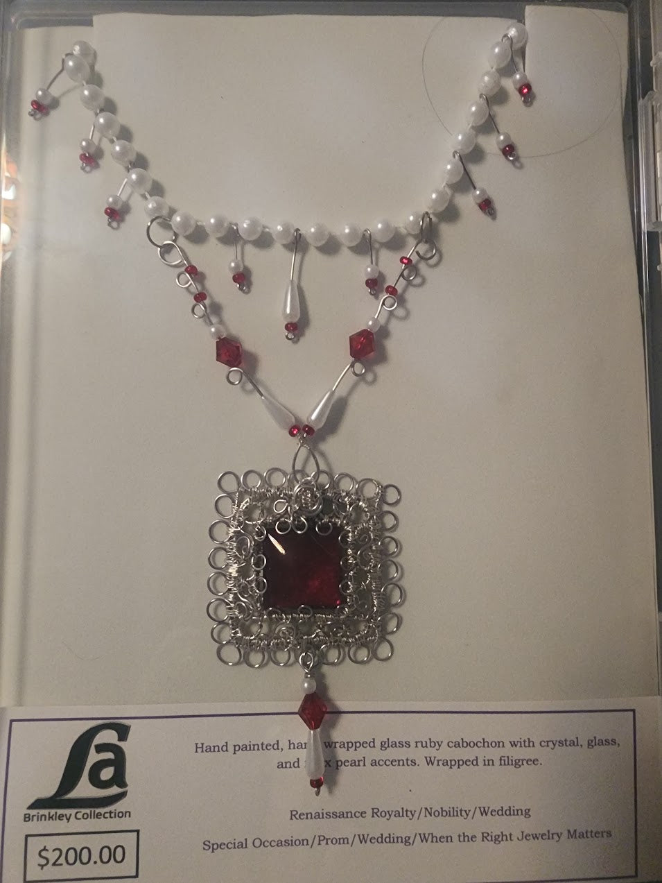 Necklace with red pendant and pearl accents on a white card with brand logo
